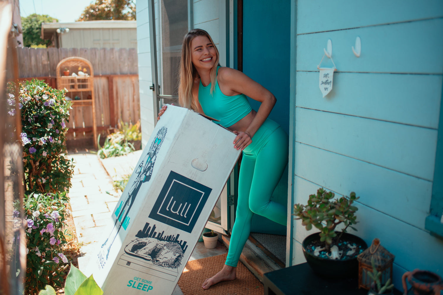 A woman in workout attire smiling while she drags a lull mattress in its box through her doorway.